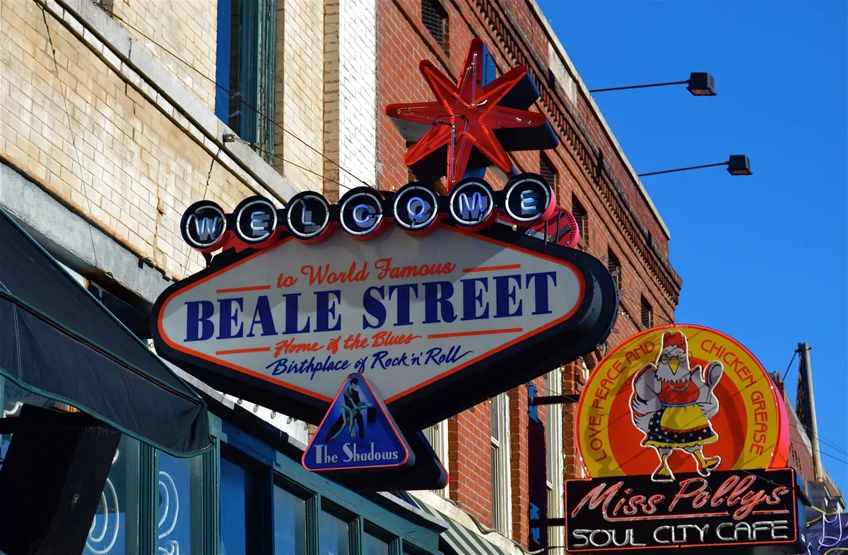 A vibrant street scene on Memphis’ iconic Beale Street, featuring the colorful 'Welcome to Beale Street' sign, inviting visitors to explore the heart of the city. The image captures bustling activity with people enjoying live music, dining at local cafes such as Miss Polly's Soul City Cafe, and soaking in the festive atmosphere under a clear sky.