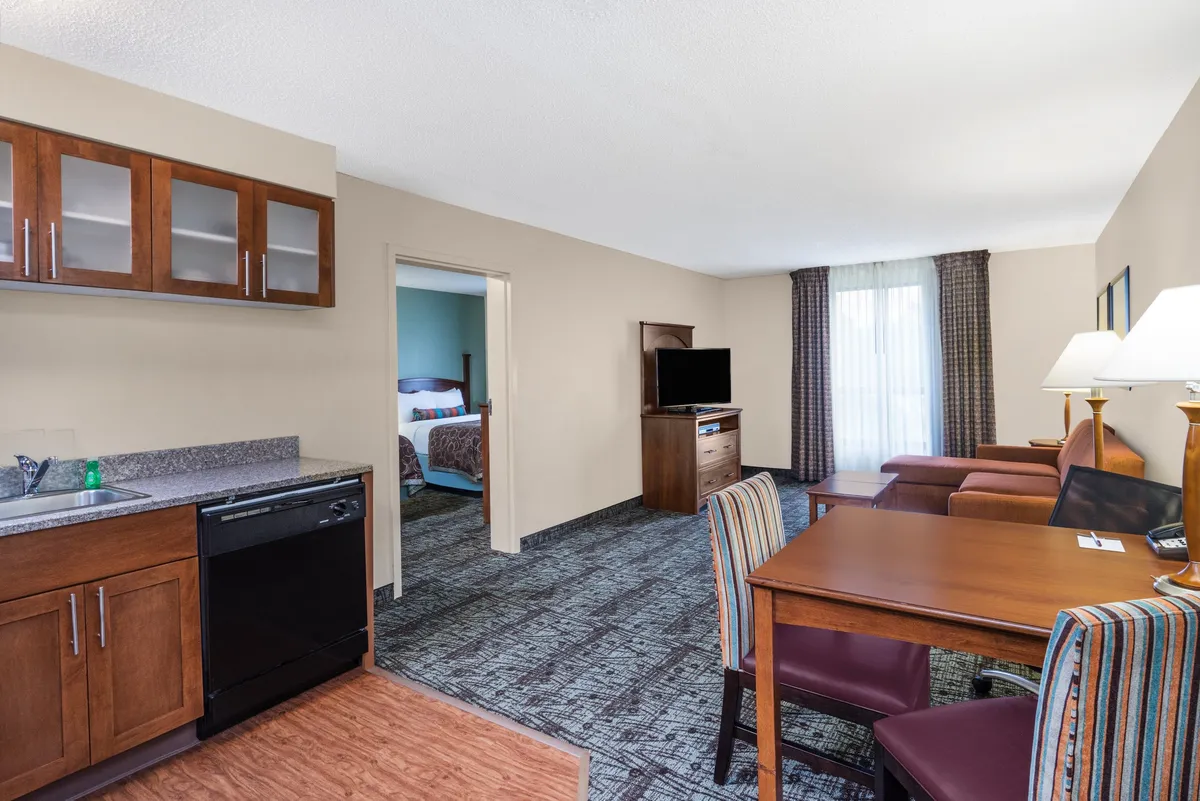 The image showcases a spacious, open-concept condo suite featuring a well-equipped kitchenette, a cozy dining area for shared meals, a comfortable living space for relaxing or watching TV, and a glimpse of the inviting bedroom entrance. The scene exudes a welcoming and functional atmosphere, perfect for both relaxation and productivity.