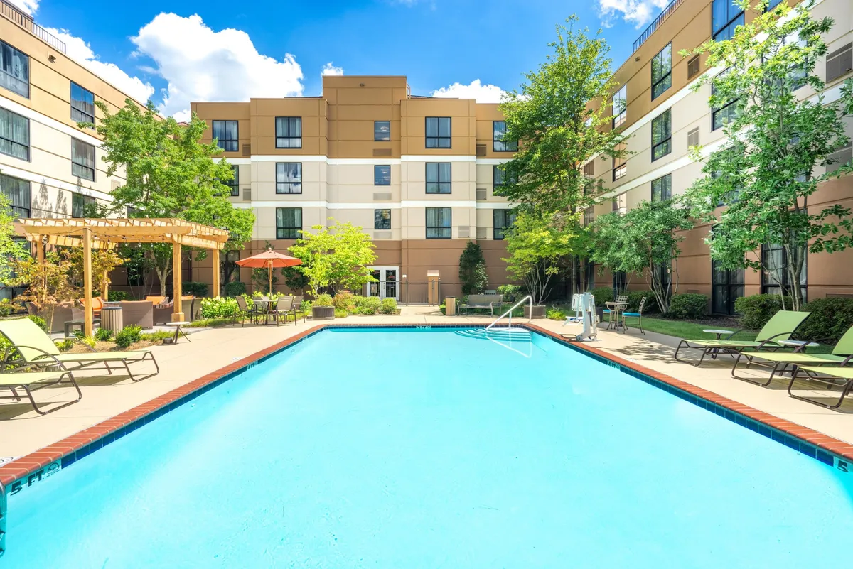 A sparkling outdoor swimming pool surrounded by inviting lounge chairs, a pergola for shade, and lush landscaped gardens. Guests are seen swimming, sunbathing, and socializing, creating a vibrant but relaxed atmosphere.