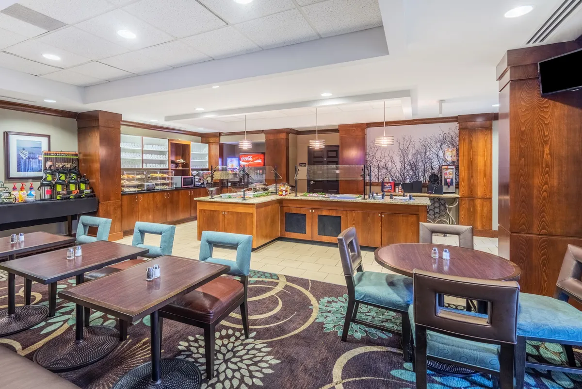 A communal dining area featuring a wooden buffet counter, neatly set tables, a vibrant carpet pattern, and shelves with glassware. The setup is ideal for casual dining, socializing, or catching up on work, offering a relaxed yet lively environment for guests to interact or find a quiet spot for productivity.