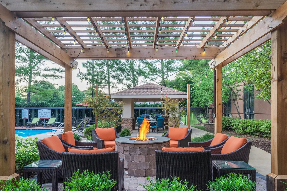 An outdoor area featuring a cozy fire pit encircled by comfortable seating and a wooden pergola, set within manicured landscaping. The scene is ideal for evening gatherings, with guests relaxing, socializing, and enjoying sunset views.